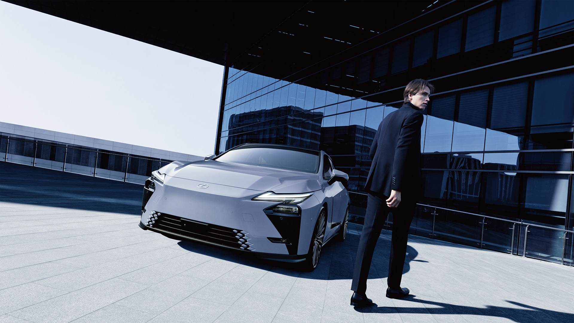 A man in a dark suit stands beside a 2026 Lexus ES All-Electric on an open plaza, with a glass-facade building reflected in the background.