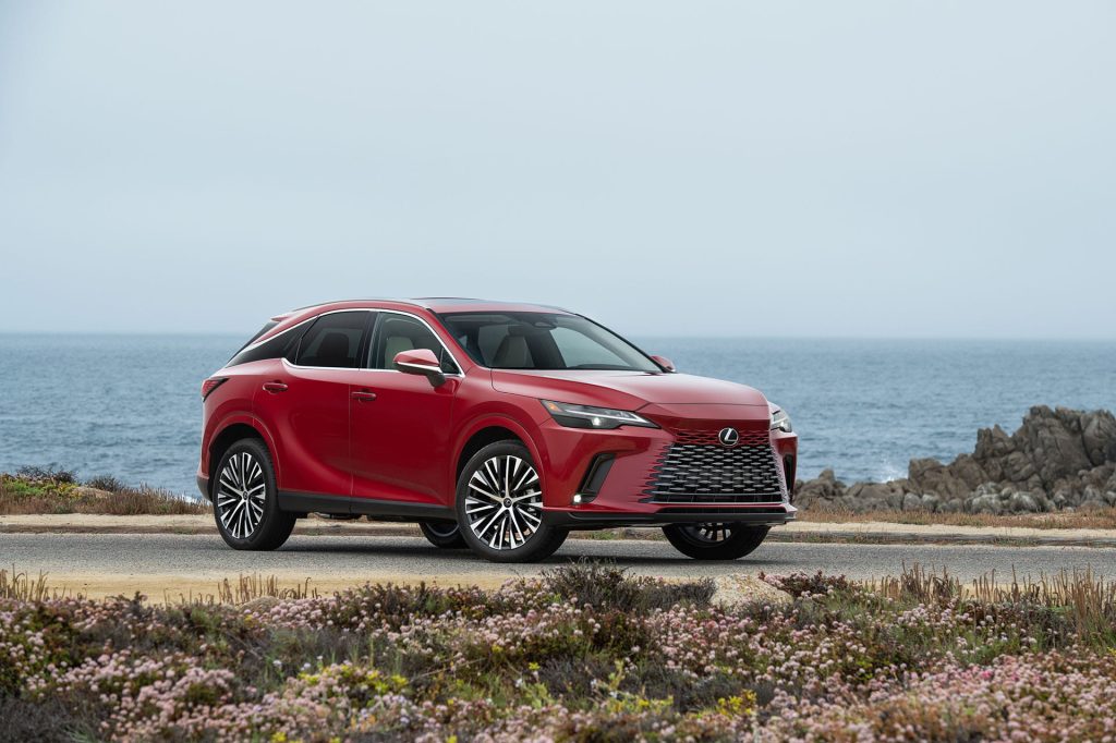 Red Lexus RX 350h luxury hybrid SUV parked by a coastal road with ocean and rocky shoreline in the background.