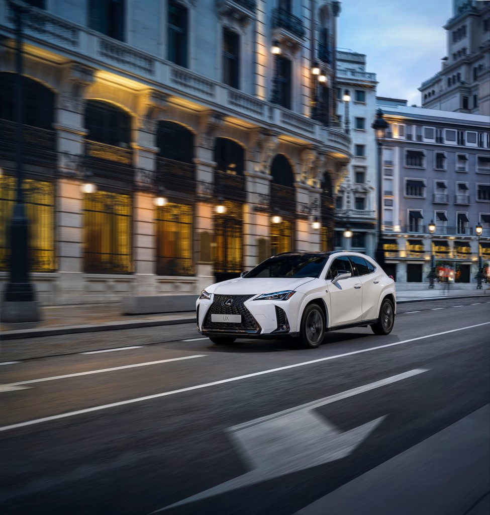 White Lexus UX compact SUV driving through a European-style city street at dusk with illuminated buildings.