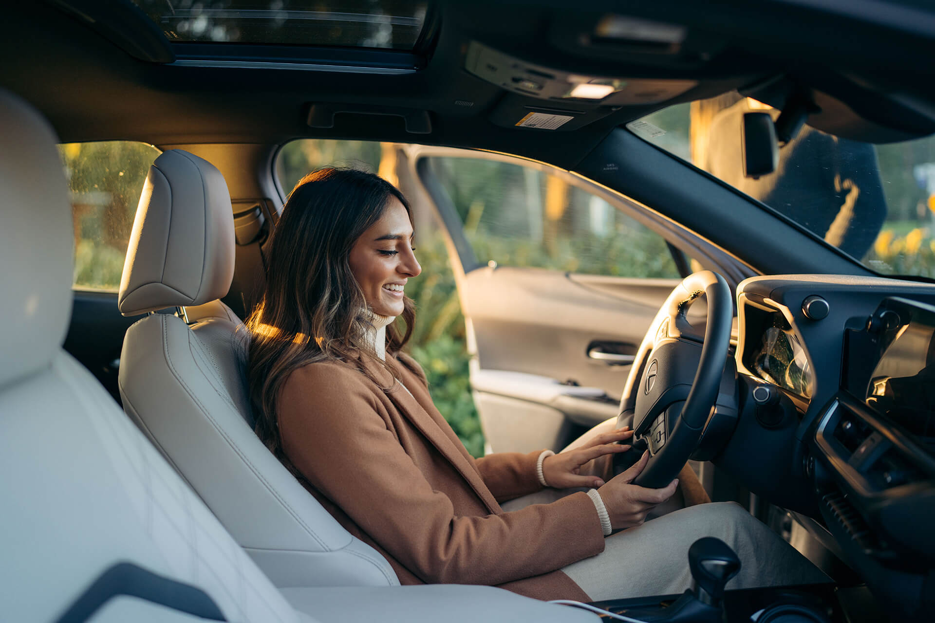 Smiling woman sitting in the driver’s seat of a modern car, adjusting the steering wheel in a bright, sunlit interior with the door open.