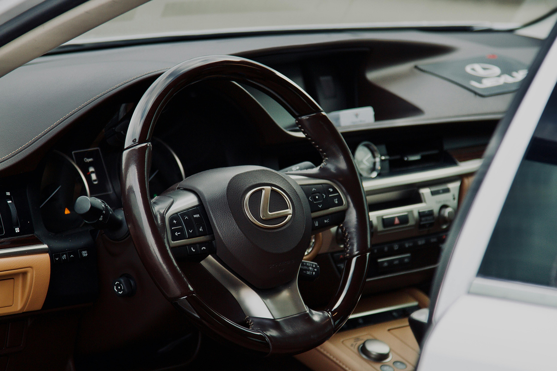 Close-up of a Lexus car interior showing a sleek black steering wheel with logo, dashboard controls, and brown leather accents, conveying luxury and sophistication.